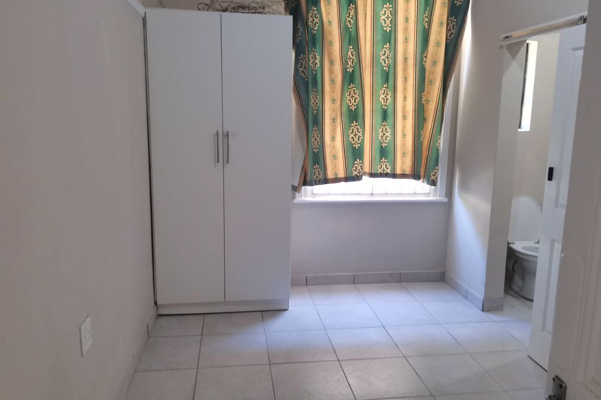 A simple bedroom featuring tiled flooring, a built-in wardrobe, and a window with security bars, offering a functional and clean living space.