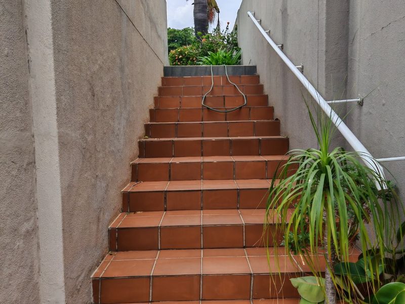 Exterior staircase featuring terracotta tiling and a white metal handrail, leading up between two boundary walls with visible plaster weathering.
