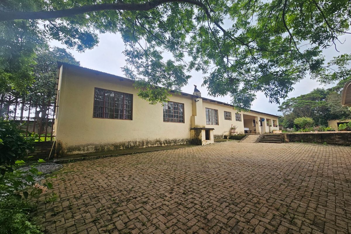 Rear exterior view of the farmhouse featuring a large paved courtyard, external chimney, and established trees surrounding the building structure.