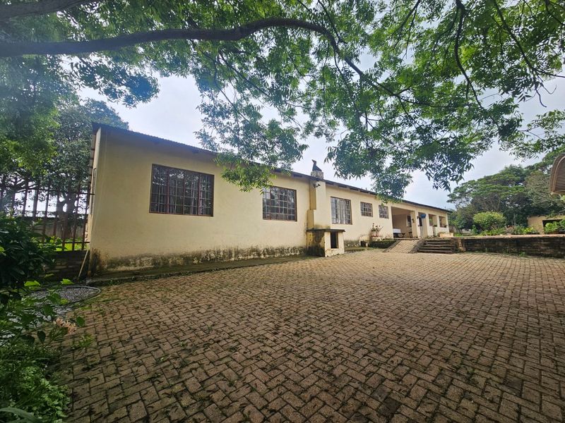 Rear exterior view of the farmhouse featuring a large paved courtyard, external chimney, and established trees surrounding the building structure.