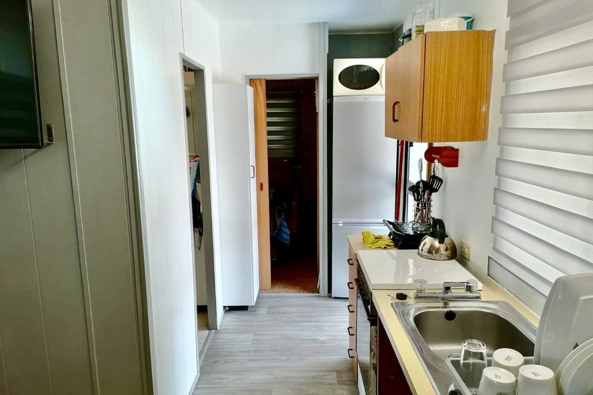 A compact galley-style kitchen featuring laminate flooring, built-in cabinetry, a stainless steel sink, and roller blinds.