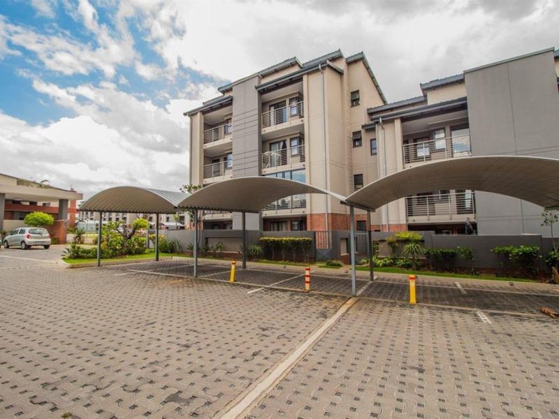 Exterior view of a modern apartment complex featuring covered parking bays, paved driveway, and multi-story residential units with balconies.