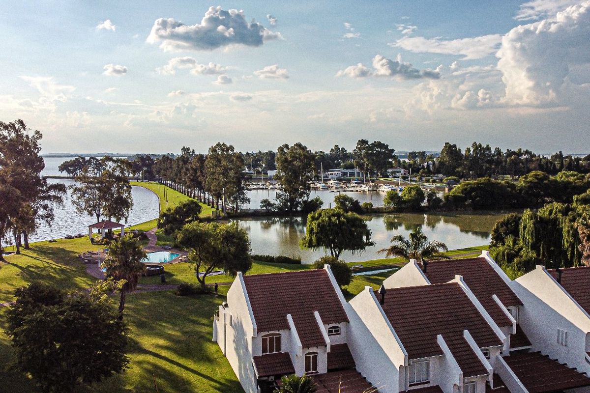 Aerial view of a residential estate featuring white townhouses with tiled roofs, expansive green lawns, a swimming pool, and scenic waterfront views.
