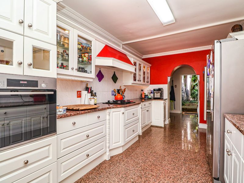 A functional kitchen featuring white shaker-style cabinetry, granite countertops, built-in oven, gas hob, and polished granite flooring, leading through an archway to the rear.