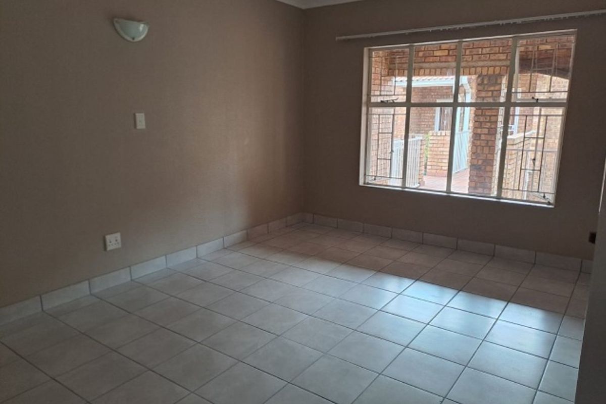 An empty living room featuring neutral tiled flooring, a wall-mounted light fixture, and a large window with security bars overlooking an exterior area.