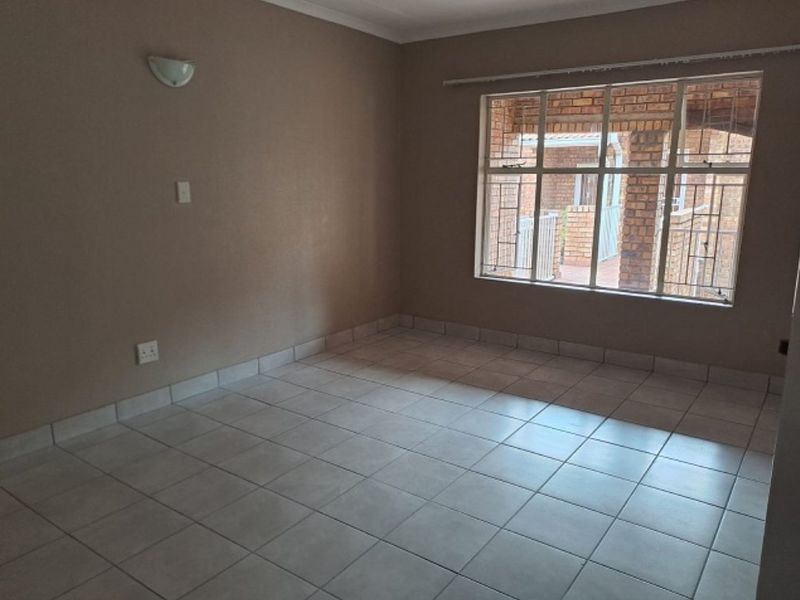 An empty living room featuring neutral tiled flooring, a wall-mounted light fixture, and a large window with security bars overlooking an exterior area.