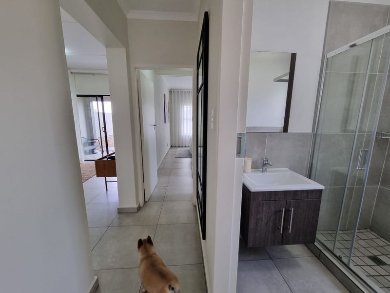 A bright hallway featuring large format floor tiles, providing access to a modern bathroom with a vanity and glass-enclosed shower.