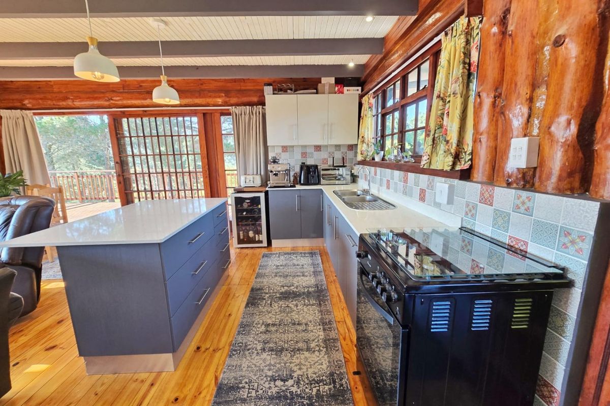 A rustic kitchen featuring timber log walls, exposed beams, a central island, modern cabinetry, and a freestanding stove, opening onto an outdoor deck.