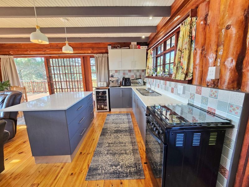 A rustic kitchen featuring timber log walls, exposed beams, a central island, modern cabinetry, and a freestanding stove, opening onto an outdoor deck.