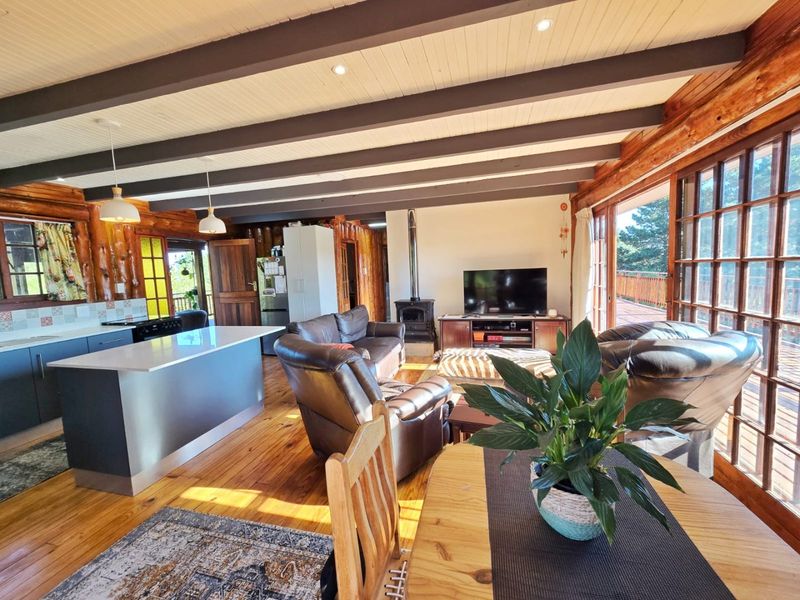 Spacious open-plan living area featuring exposed log walls, hardwood floors, a modern kitchen island, and a cozy wood-burning stove with access to a deck.