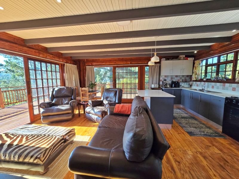 Spacious open-plan living and kitchen area featuring exposed timber beams, warm wood flooring, and large glass doors opening onto a deck with scenic views.