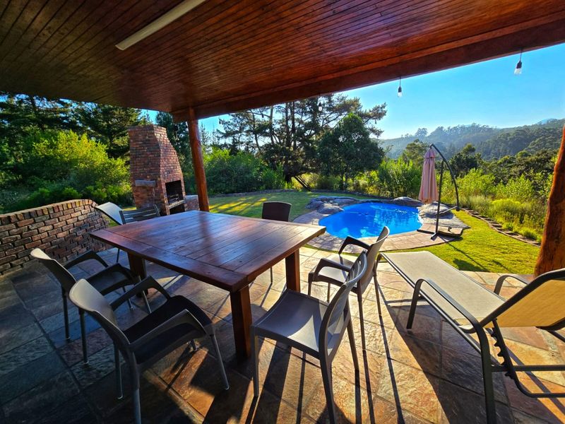 Covered outdoor patio featuring a built-in brick braai, stone flooring, and timber ceiling, overlooking a private swimming pool and landscaped garden.