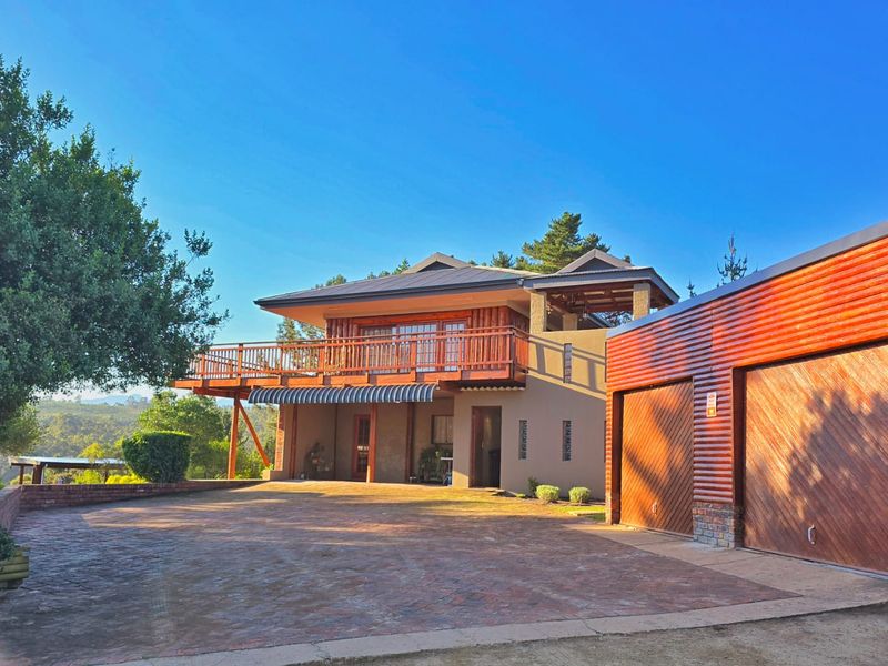 Front view of a farm-style property featuring a double garage, brick paved driveway, and a raised timber deck with scenic surroundings.