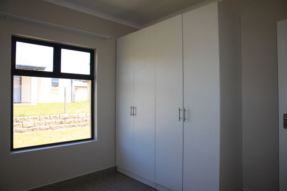 A clean, empty bedroom featuring modern white built-in cupboards, tiled flooring, and a large window providing natural light.