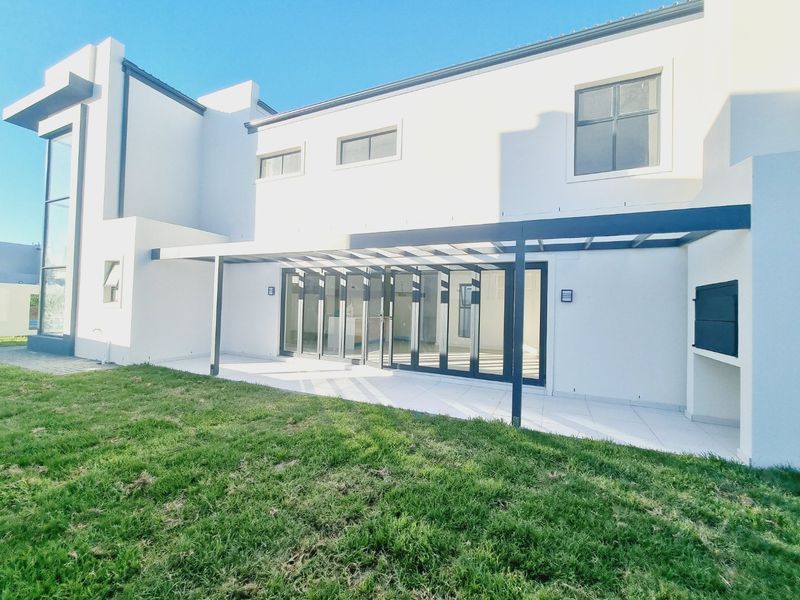 Rear view of a modern home featuring a covered patio with built-in braai, large sliding glass doors, and a well-maintained lawn.