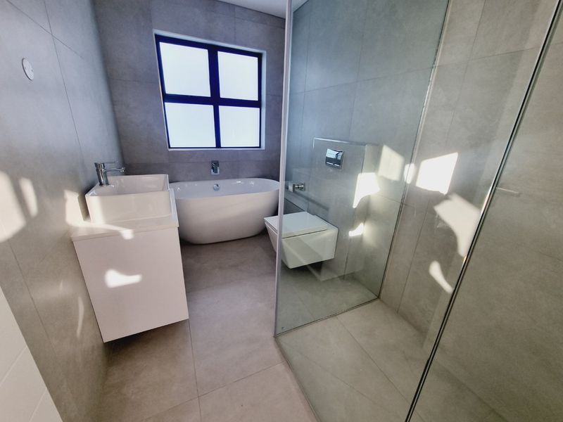 Modern bathroom featuring a freestanding bathtub, wall-mounted toilet, glass-enclosed shower, and contemporary vanity with neutral grey tiling throughout.
