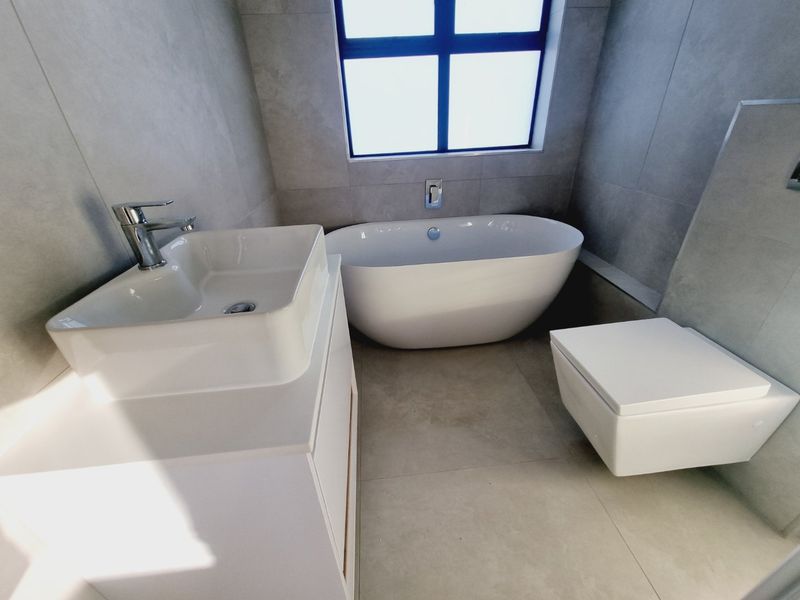 Modern bathroom featuring a sleek freestanding bathtub, wall-mounted toilet, and a contemporary vanity basin with integrated cabinetry, finished with neutral floor-to-ceiling tiles.