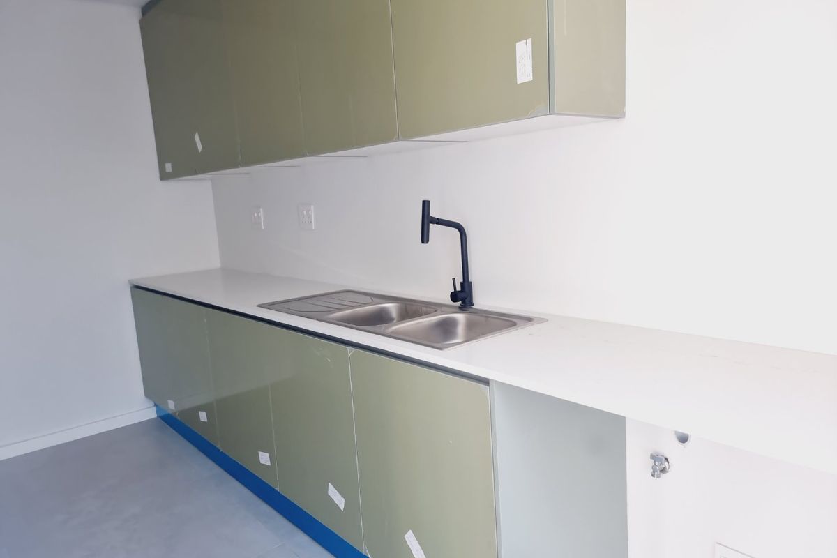 A modern, minimalist kitchen featuring sage green cabinetry, a stainless steel double sink with a matte black tap, and clean white countertops.
