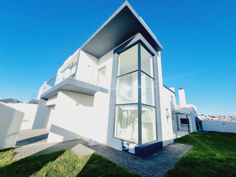 Modern double-storey residence featuring a striking double-height glass facade, paved patio area, and built-in outdoor braai, set within a well-maintained garden.