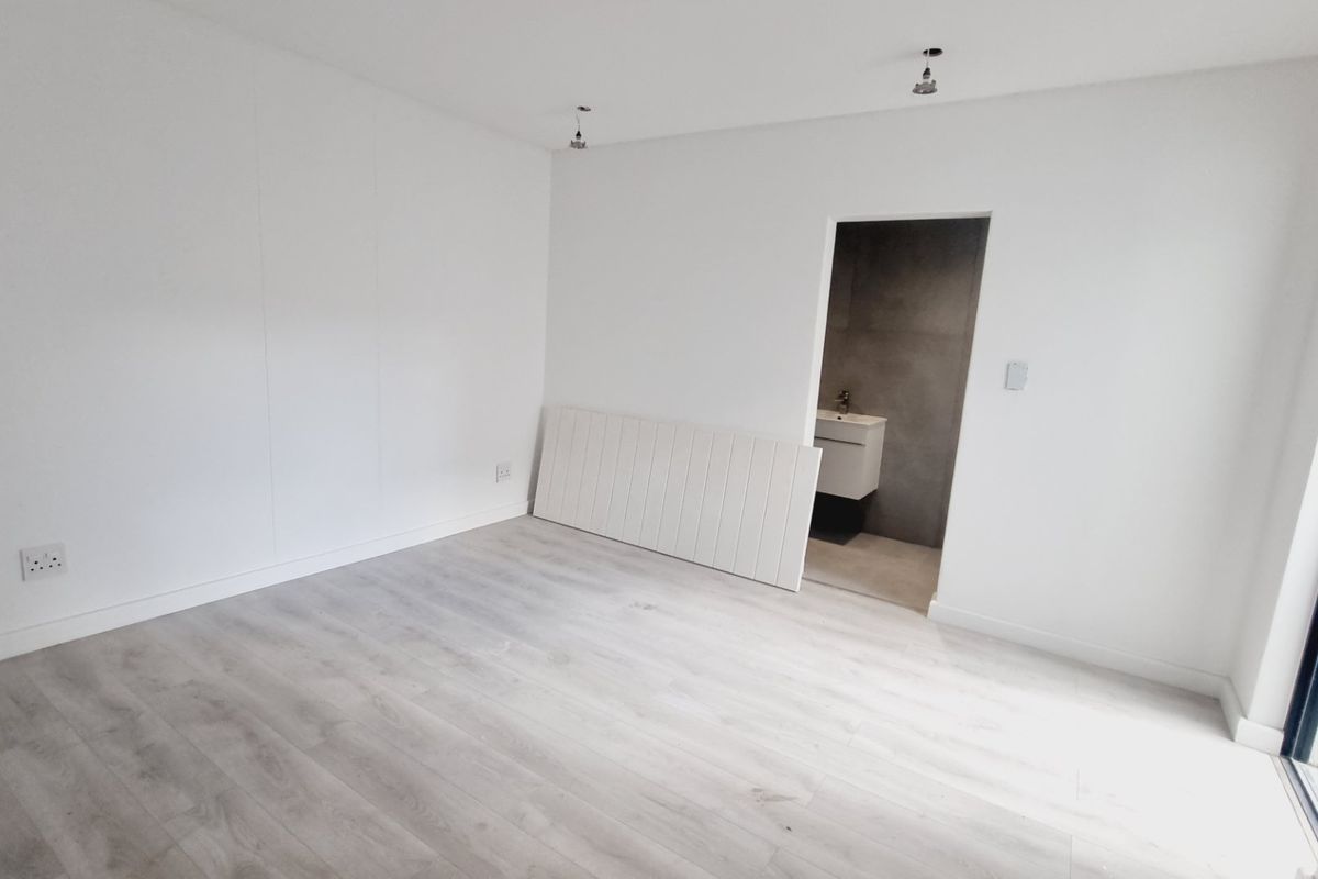 A bright, empty bedroom featuring light-toned laminate flooring, neutral walls, and direct access to an en-suite bathroom with modern finishes.