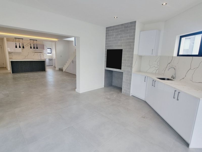 A modern open-plan living and kitchen area featuring sleek white cabinetry, stone-look countertops, a built-in fireplace with grey brick detailing, and large format floor tiles.