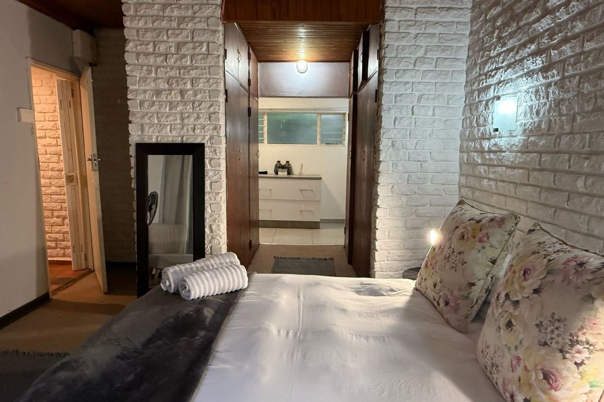 A bedroom featuring exposed white brick walls and a timber ceiling, with built-in cupboards and an adjoining space visible through the central opening.