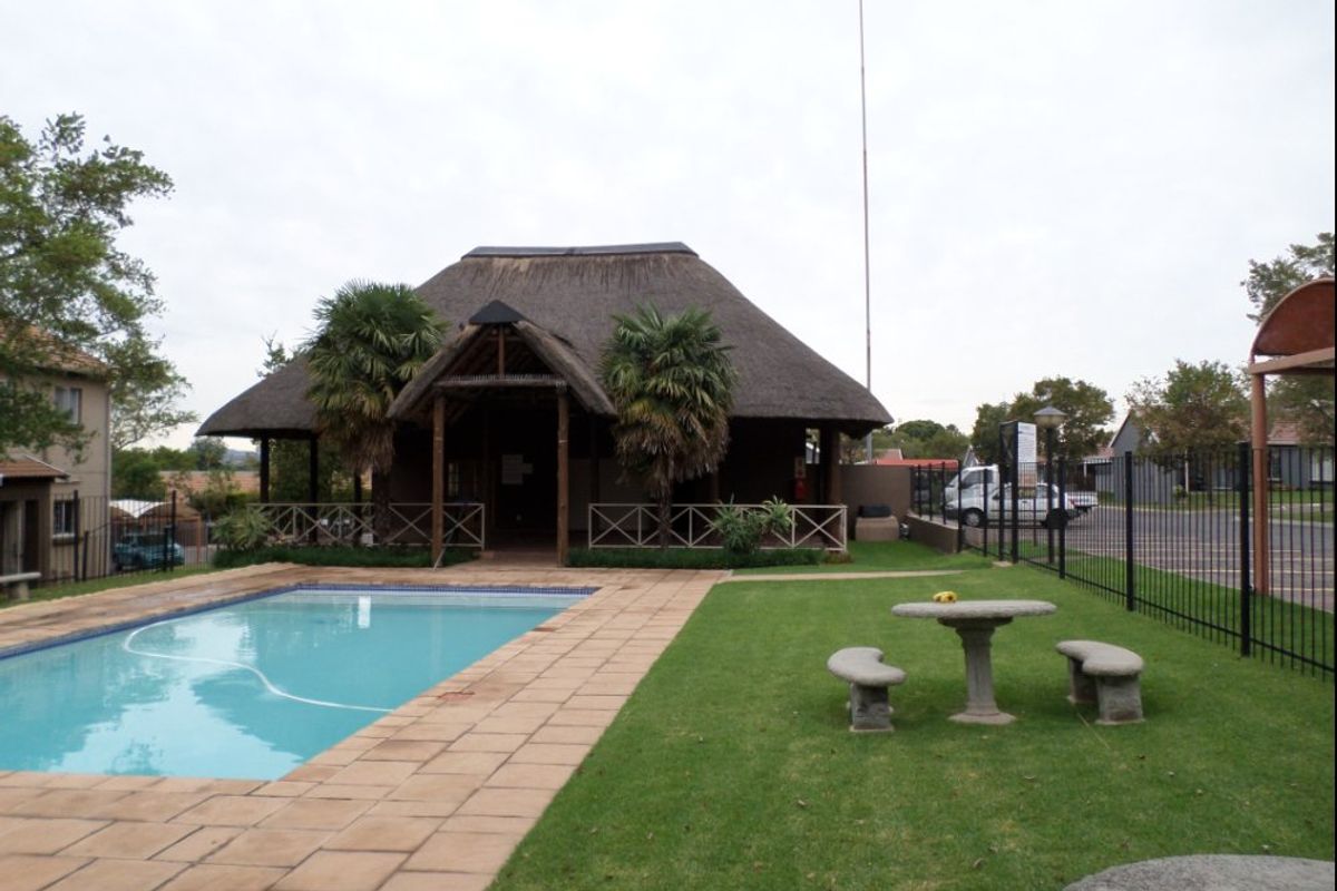 Communal pool area featuring a swimming pool, paved deck, and a thatched roof clubhouse set within a landscaped garden.