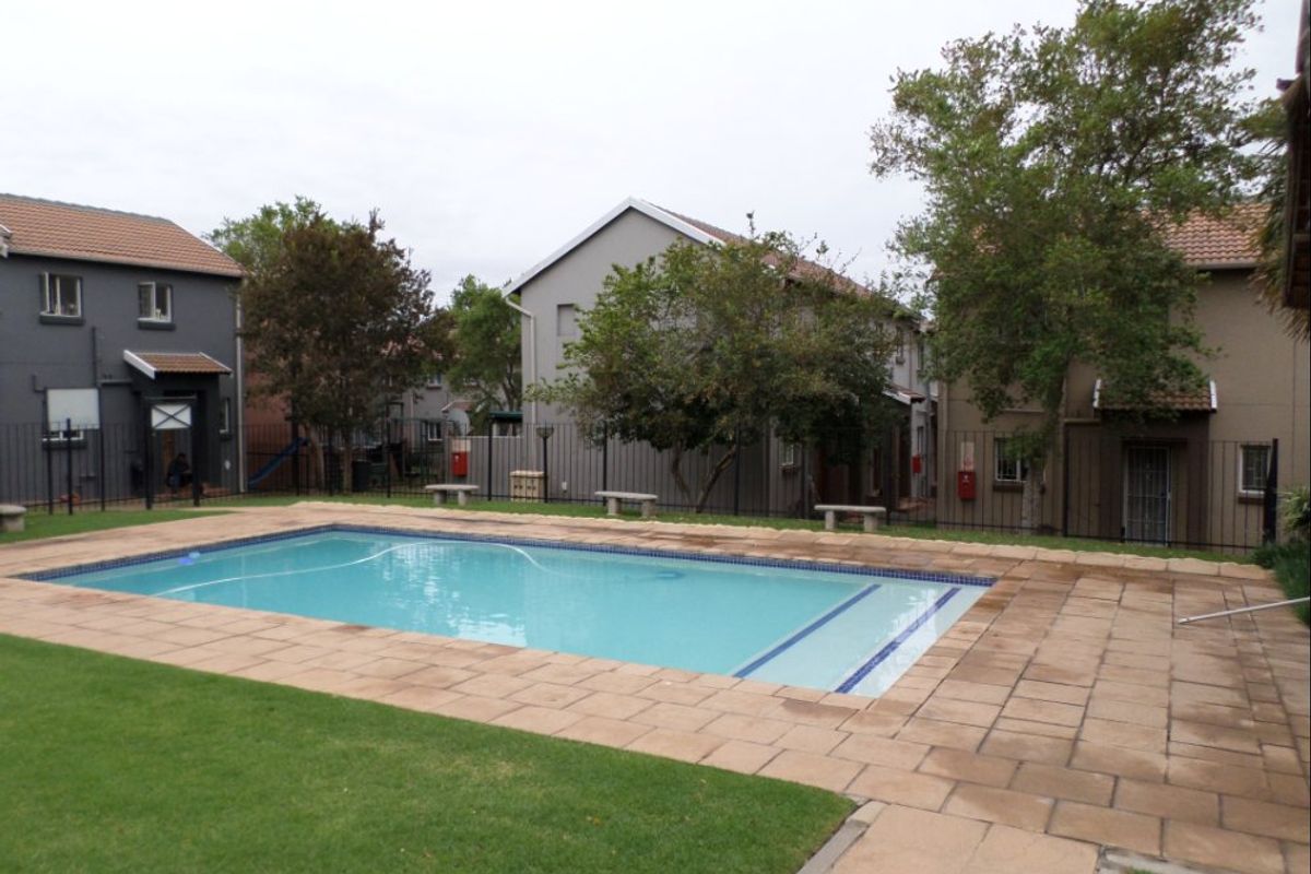 Communal swimming pool area featuring a paved deck, surrounding lawn, and secure metal perimeter fencing within an apartment complex setting.