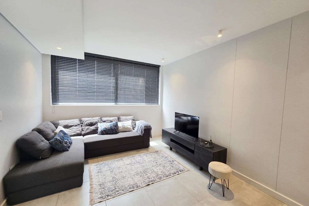A modern living room featuring neutral tiled flooring, recessed ceiling lighting, and large windows fitted with dark venetian blinds.