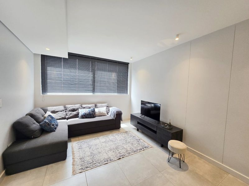 A modern living room featuring neutral tiled flooring, recessed ceiling lighting, and large windows fitted with dark venetian blinds.