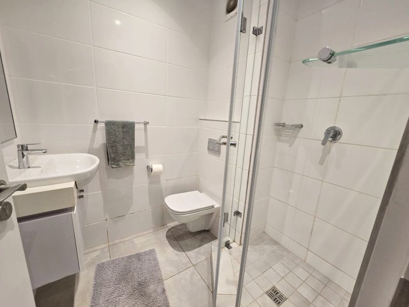 Compact modern bathroom featuring a wall-mounted toilet, vanity basin, and a glass-enclosed shower with white tiled finishes.