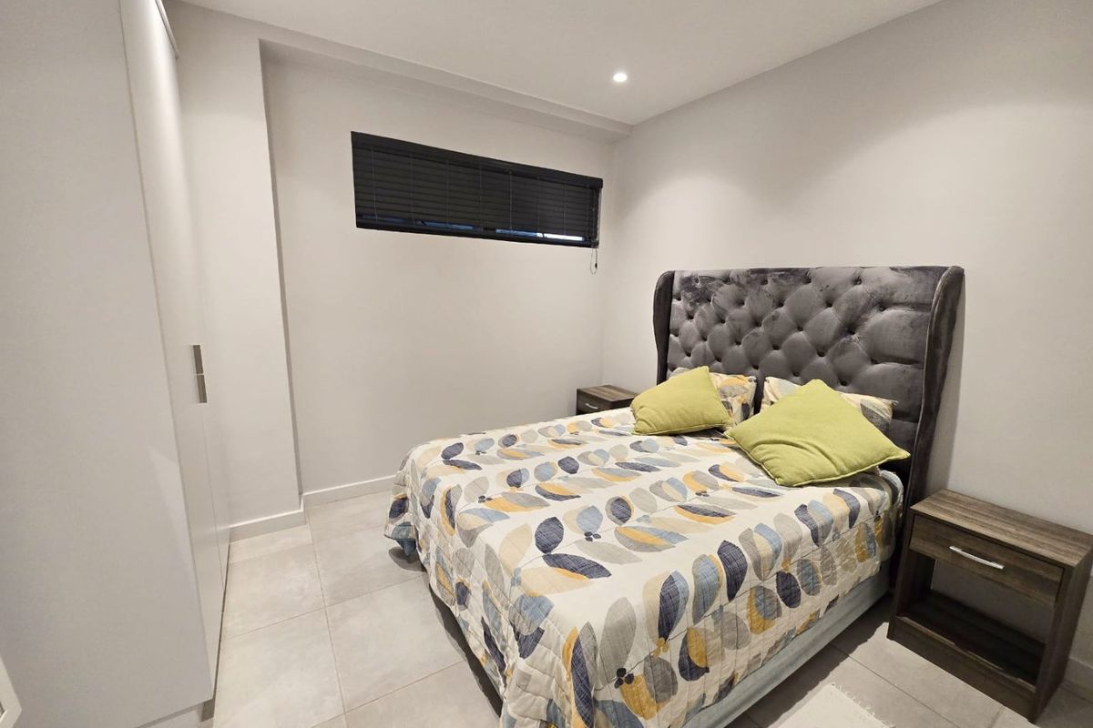 A modern bedroom featuring neutral tiled flooring, built-in wardrobe storage, recessed downlighting, and a high-set horizontal window with blinds.