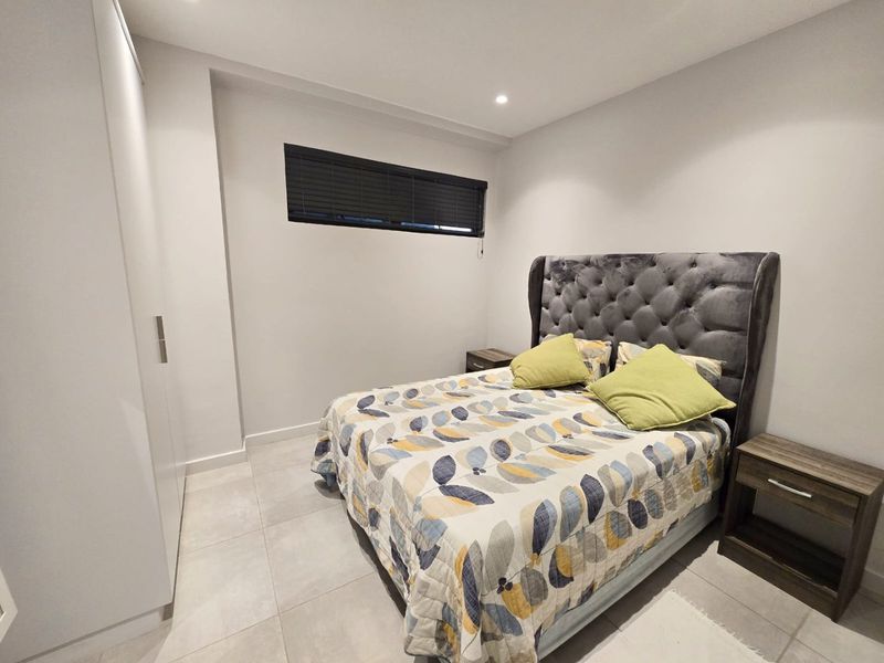 A modern bedroom featuring neutral tiled flooring, built-in wardrobe storage, recessed downlighting, and a high-set horizontal window with blinds.
