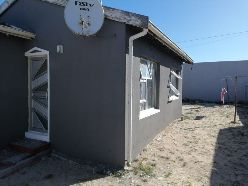 Rear exterior view of the house featuring grey painted walls, aluminium window frames, and a security door, with an unpaved yard area.