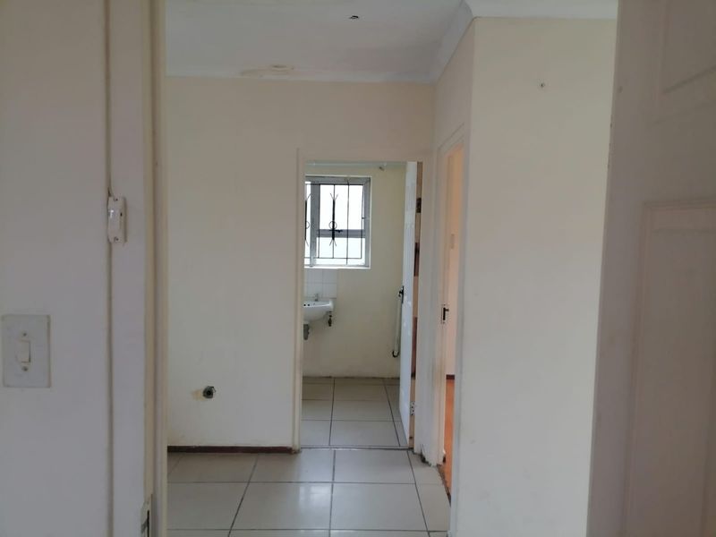 A vacant hallway with tiled flooring leading to a small bathroom area featuring a wall-mounted basin and security bars on the window.