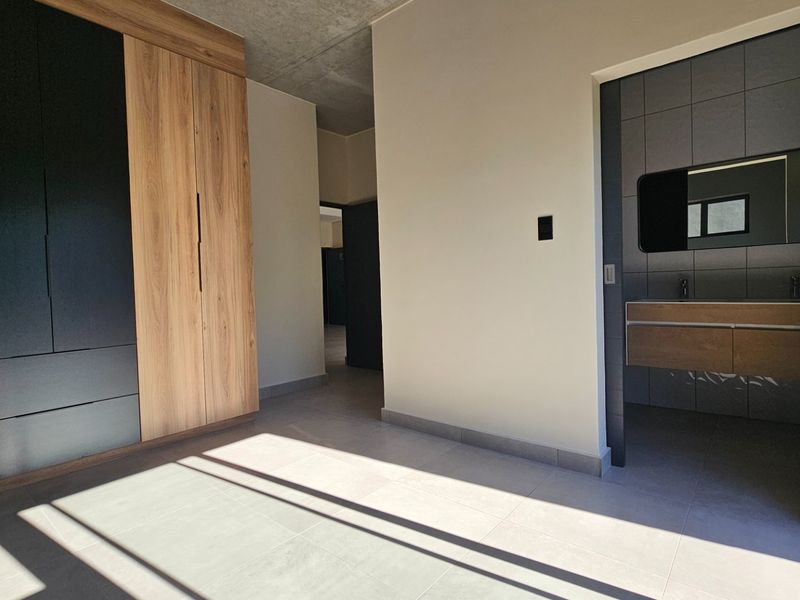 Modern bedroom featuring built-in cupboards, tiled flooring, and an exposed concrete ceiling, with an adjoining en-suite bathroom equipped with a floating vanity.