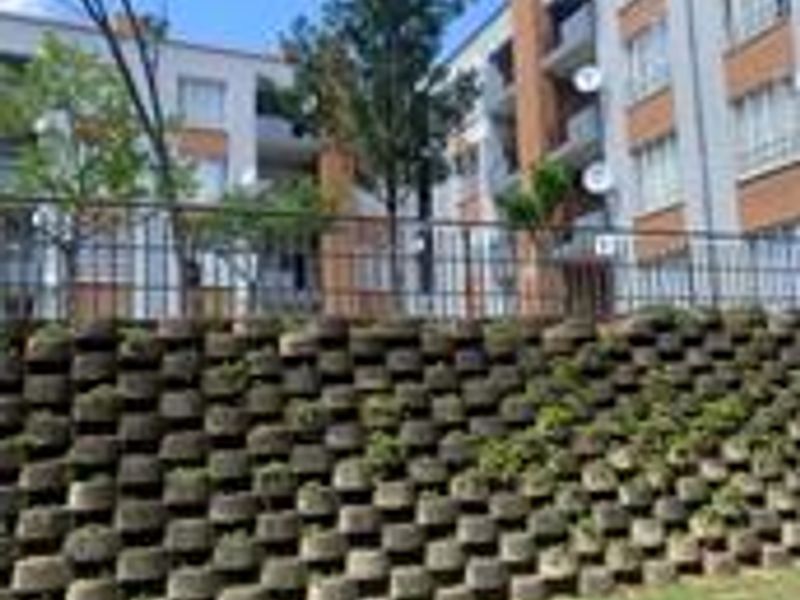 Exterior view of the apartment complex featuring a tiered concrete retaining wall, landscaped lawn, and private balconies with metal railings.