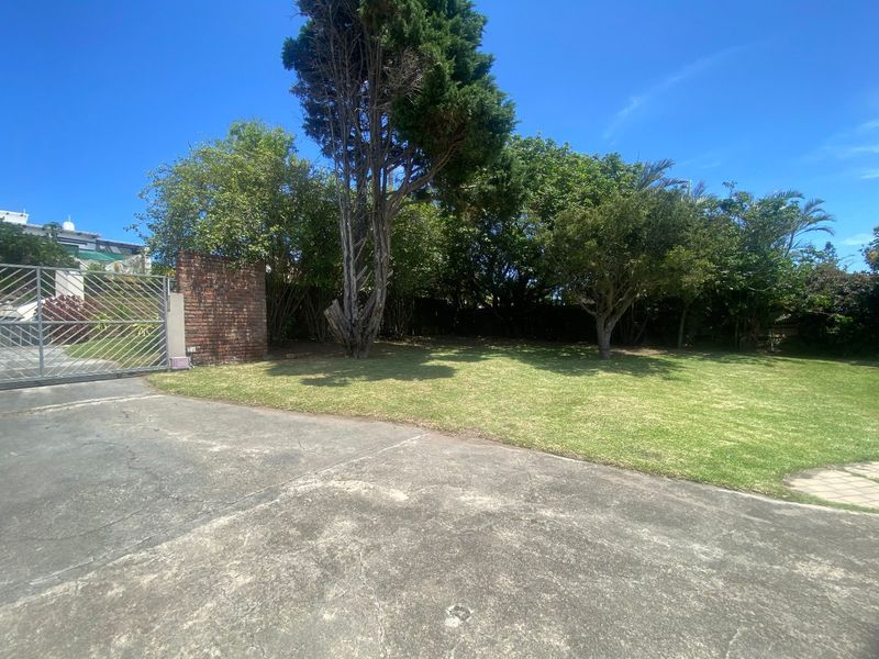A front exterior view featuring a concrete driveway, mature trees, a lawn, and a brick boundary wall with a metal security gate.