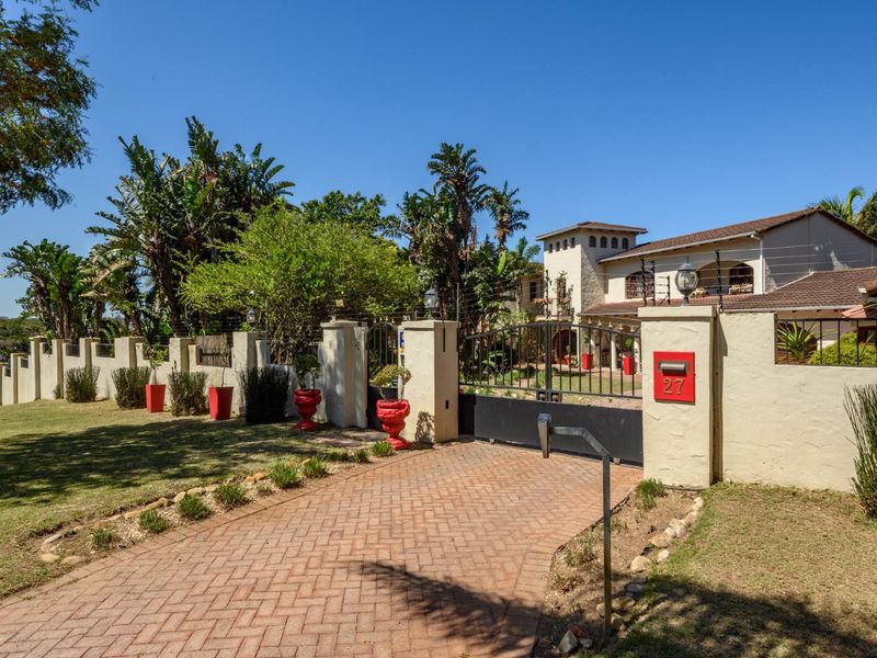 Front view of the property featuring a brick paved driveway, secure metal gate, perimeter wall with electric fencing, and established garden landscaping.