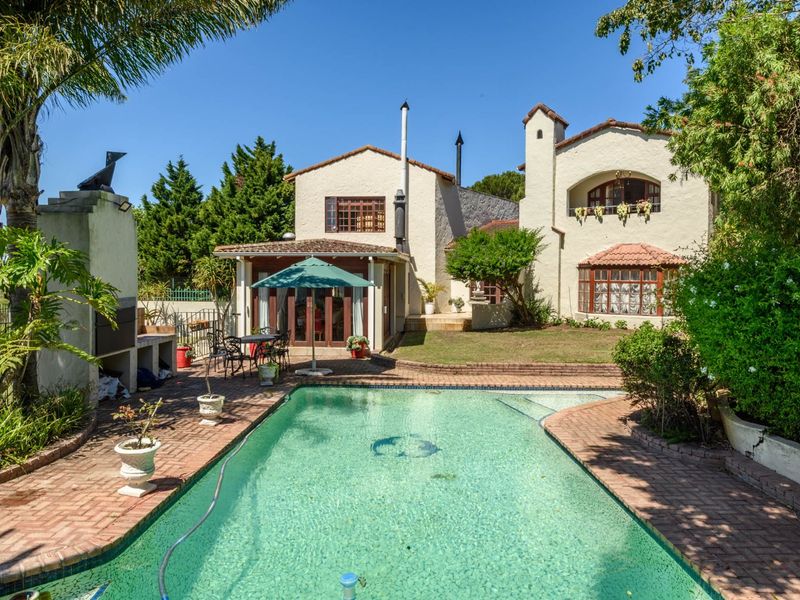 Rear view of the property featuring a swimming pool, built-in braai area, and established garden with traditional architectural styling.