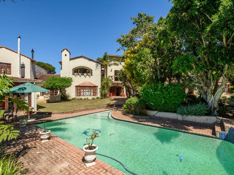 A spacious backyard featuring a swimming pool, brick paved surround, mature landscaping, and a multi-story residence with a balcony.