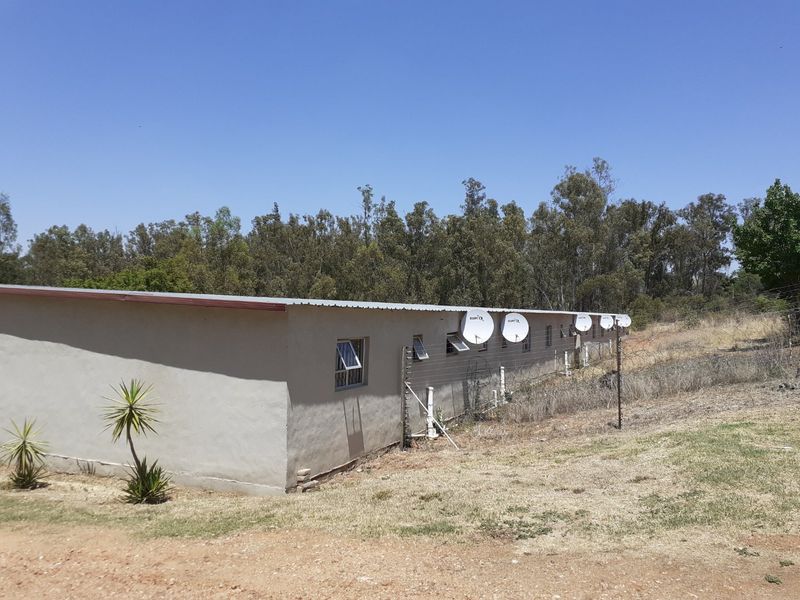 Rear view of a single-story farm building featuring plastered walls, a metal roof, and multiple satellite dishes installed along the exterior wall.