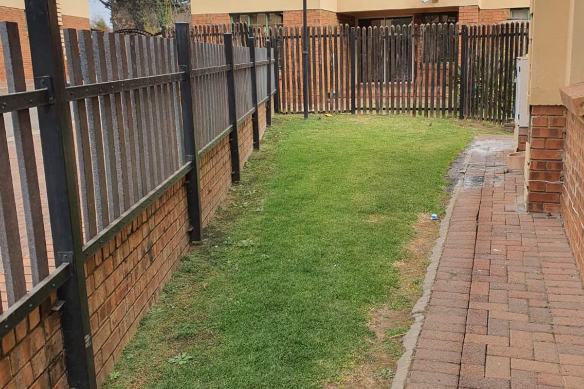 A narrow rear garden area featuring a brick-paved walkway, a small lawn, and metal perimeter fencing between townhouse units.