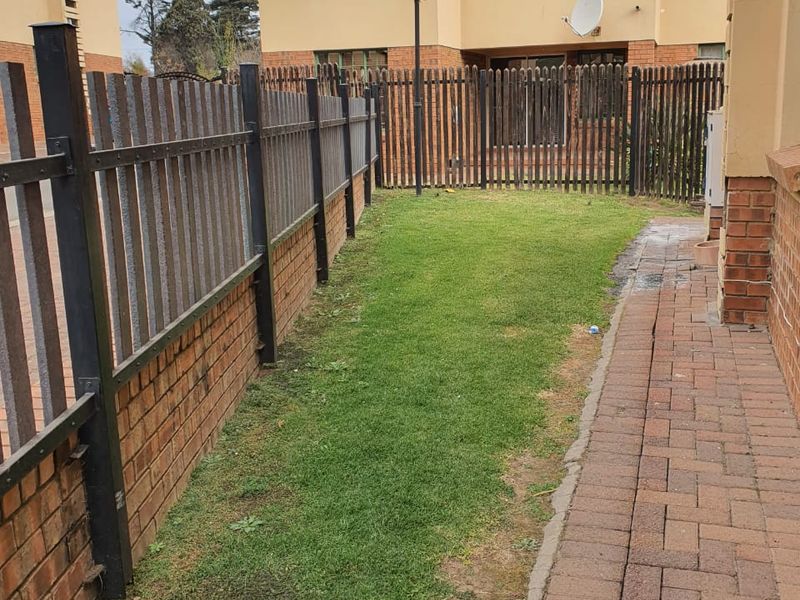 A narrow rear garden area featuring a brick-paved walkway, a small lawn, and metal perimeter fencing between townhouse units.