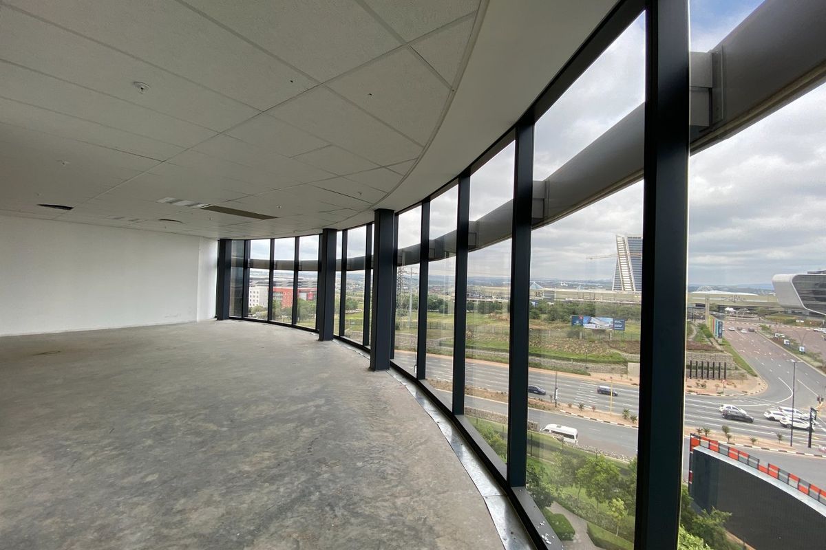 Spacious open-plan office area featuring expansive floor-to-ceiling curved windows with panoramic views, suspended ceiling, and unfinished concrete flooring.