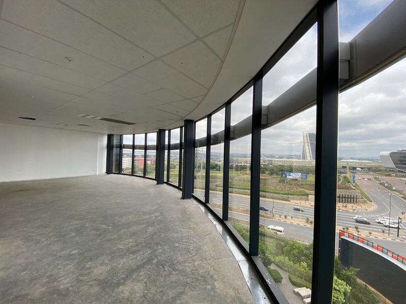 Spacious open-plan office area featuring expansive floor-to-ceiling curved windows with panoramic views, suspended ceiling, and unfinished concrete flooring.