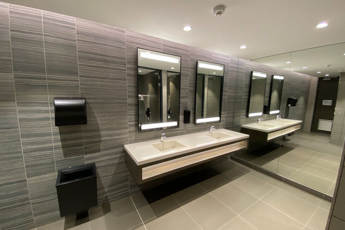 Modern commercial bathroom featuring a double vanity with integrated sinks, large wall-mounted mirrors, and contemporary grey tiled finishes.