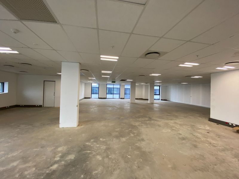 A large, open-plan commercial office space featuring a suspended ceiling, recessed lighting, structural columns, and large windows, currently in a shell condition.