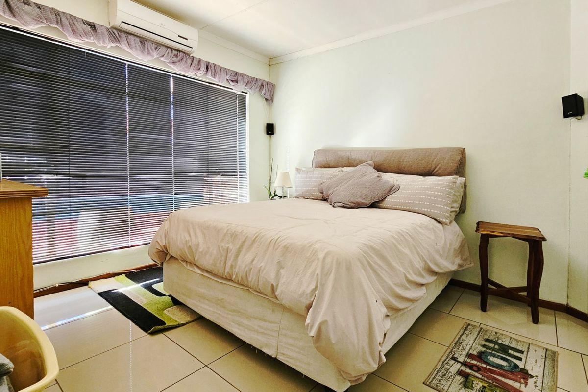 A bedroom featuring tiled flooring, a wall-mounted air conditioning unit, and large windows with fitted blinds.