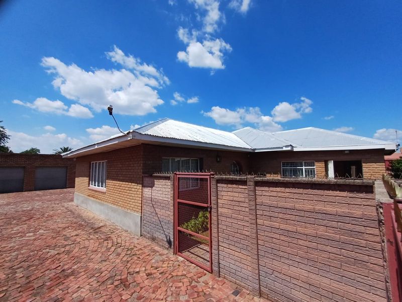 Front view of a single-story brick house featuring a paved driveway, secure boundary wall with metal gate, and an attached garage.